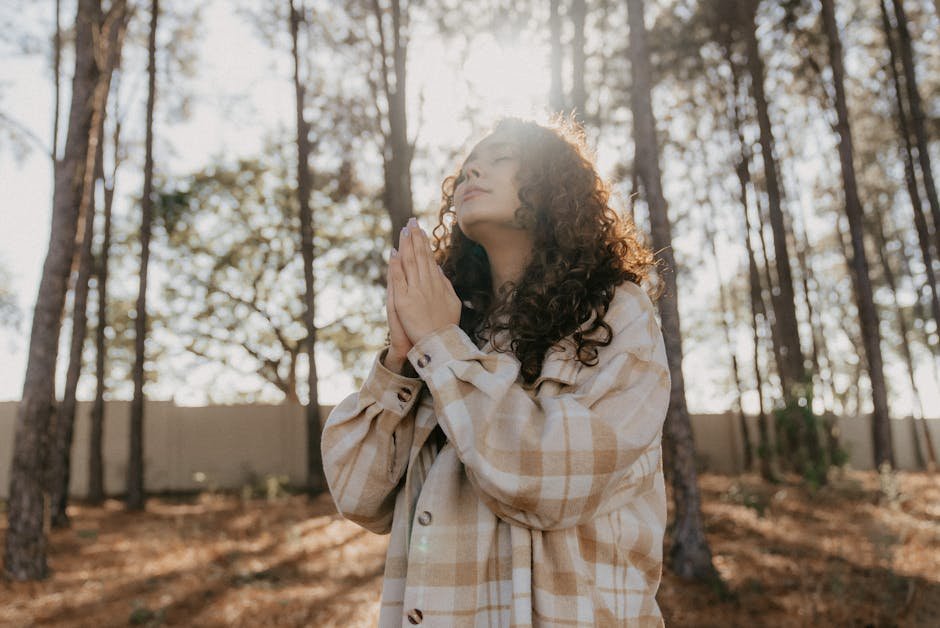 A woman in a plaid jacket stands in a sunlit forest, hands clasped in prayer.